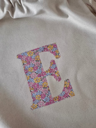 A cream canvas drawstring backpack with a personalised Liberty floral initial sewn on the front