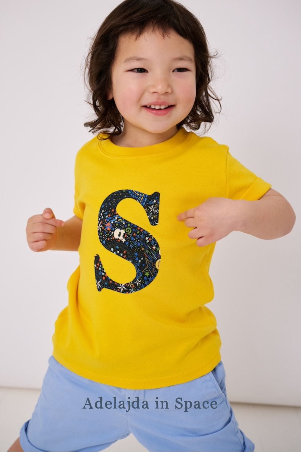a smiling 3 year old boy wearing a personalised kids yellow t-shirt with an initial cut from adelaide in space liberty  print on the front