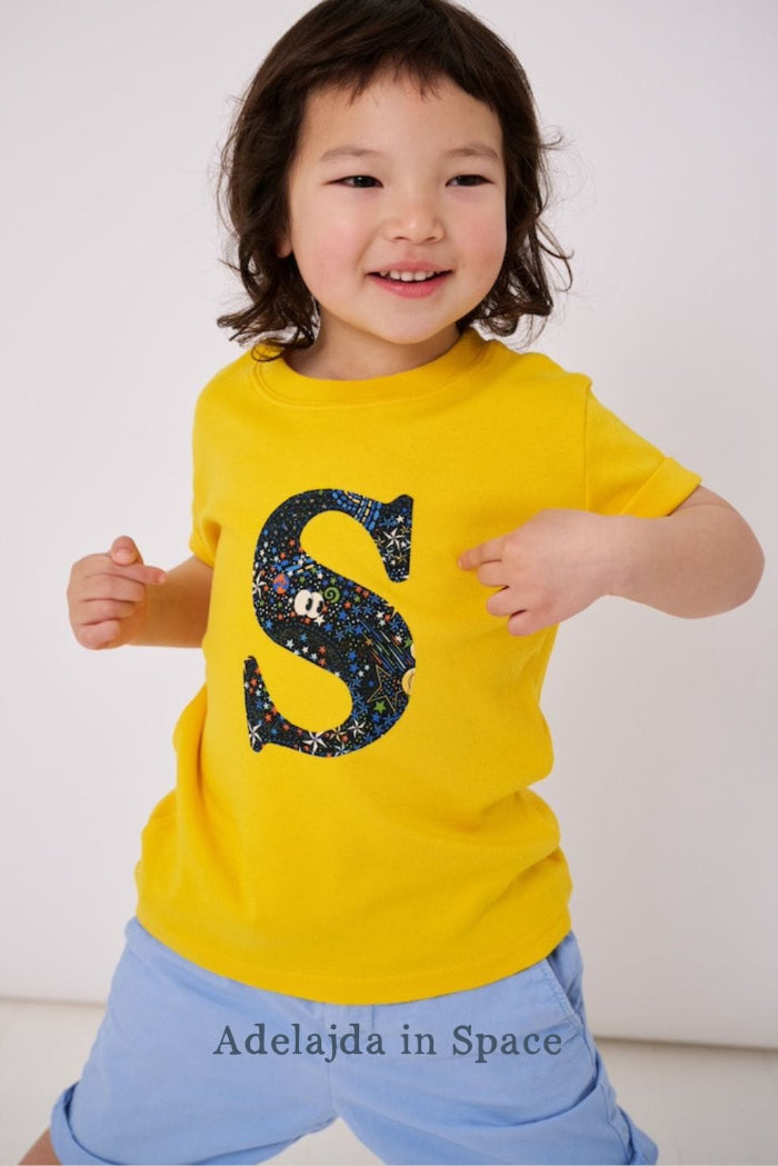 a smiling 3 year old boy wearing a personalised kids yellow t-shirt with an initial cut from adelaide in space liberty  print on the front