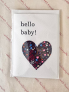 A square white greeting card with the words 'hello baby!' in black text and a heart-shaped pattern of colorful stars on the front, photographed in a clear plastic sleeve on a beige patterned surface.