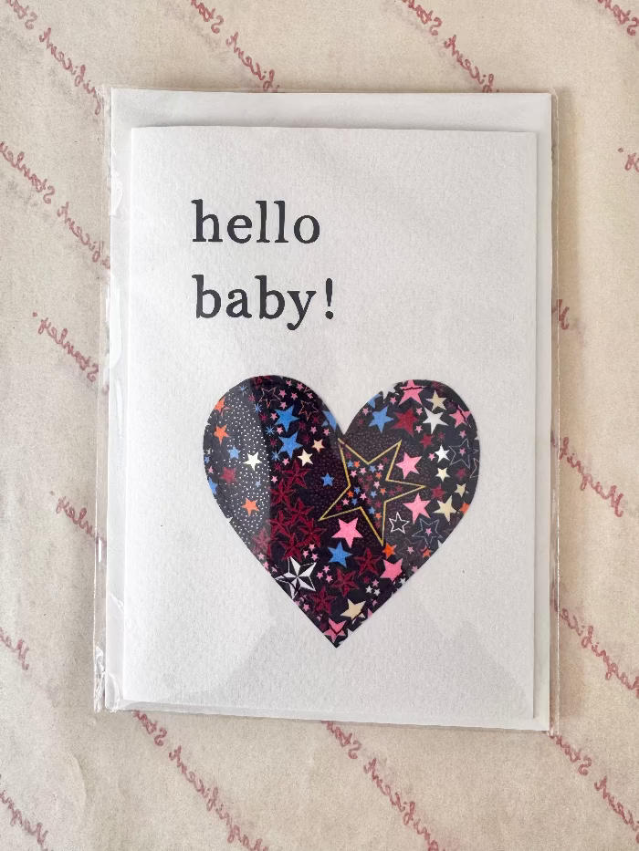 A square white greeting card with the words 'hello baby!' in black text and a heart-shaped pattern of colorful stars on the front, photographed in a clear plastic sleeve on a beige patterned surface.