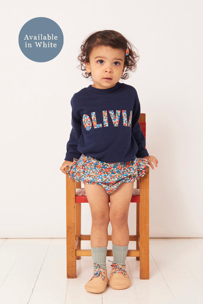 Child wearing a navy sweatshirt with 'OLIVIA' printed on it, sitting on a chair against a white background.