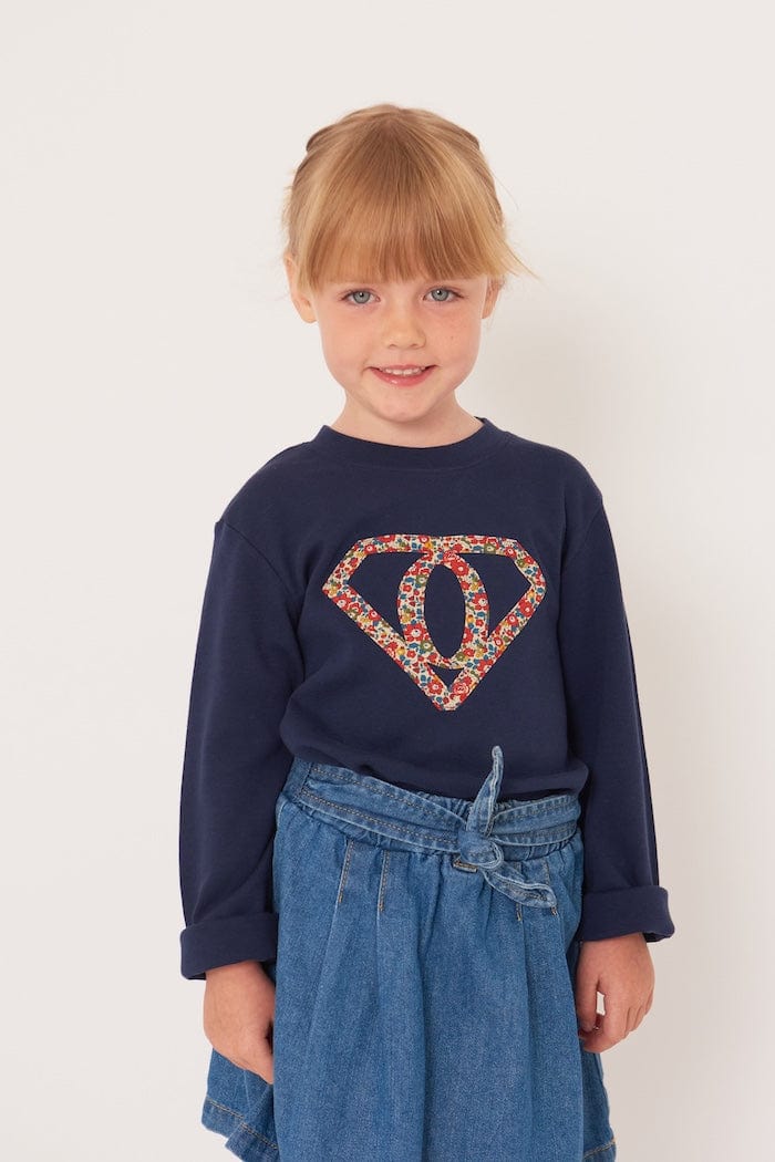 A 5 year old girl wearing a personalised  navy t-shirt with a superhero motif initial made from floral Liberty print fabric.