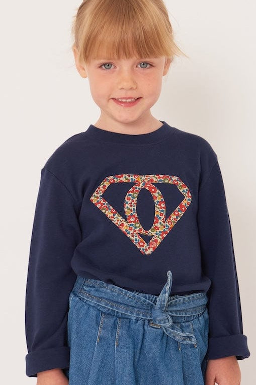 A 5 year old girl wearing a personalised  navy t-shirt with a superhero motif initial made from floral Liberty print fabric.