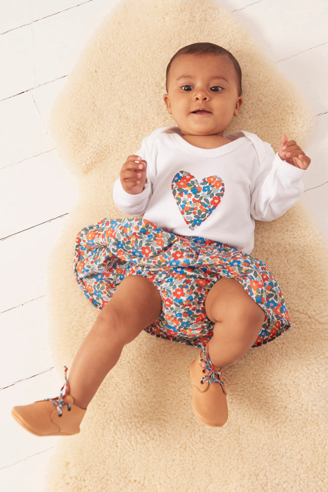 Baby wearing a white  bodysuit with a colourful  liberty print heart design and a floral skirt on a light wooden floor.