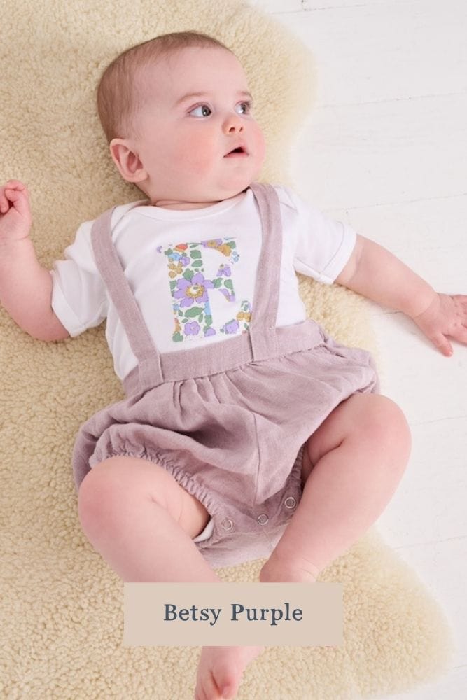 A smiling newborn baby wearing a personalised white bodysuit with an initial on the front cut from betsy purple Liberty floral fabric.