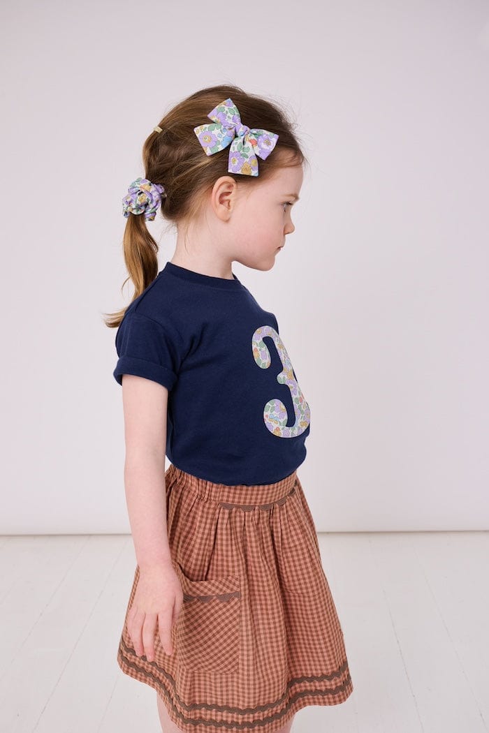 a 3 year old girl wearing a navy birthday t-shirt with a number 3 cut from liberty betsy purple floral print on the front and a matching floral scrunchie.