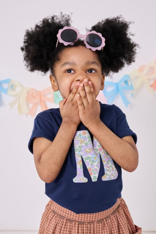 a 3 year old girl wearing a personalised kids navy t-shirt with an initial cut from betsy purple liberty floral print on the front

