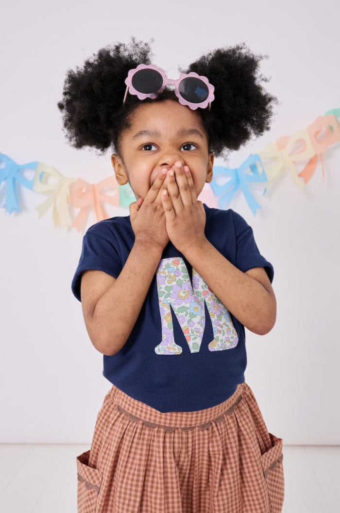 a 3 year old girl wearing a personalised kids navy t-shirt with an initial cut from betsy purple liberty floral print on the front
