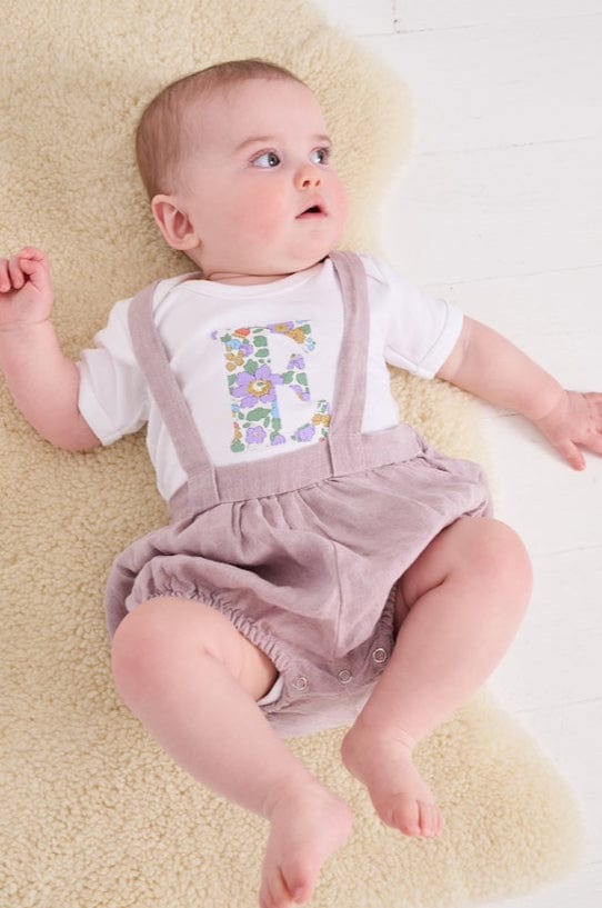 A smiling newborn baby wearing a personalised white bodysuit with an initial on the front cut from betsy purple Liberty floral fabric.