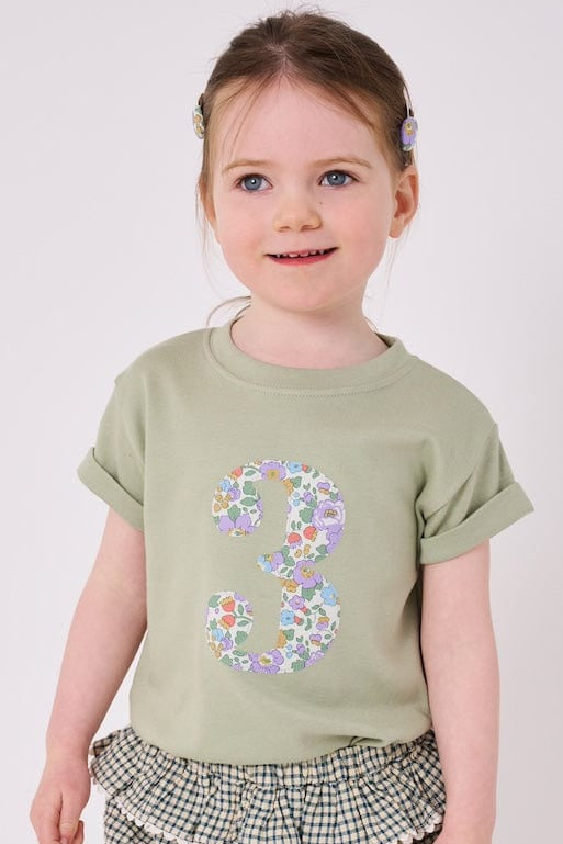 a 3 year old girl  wearing a birthday number 3 kids sage green t-shirt with a number cut from betsy purple liberty print on the front.