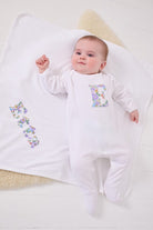 a happy baby wearing a cotton baby romper with her initial on the front in betsy purple floral liberty print. she is laying on a  cotton blanket with her name sewn on.