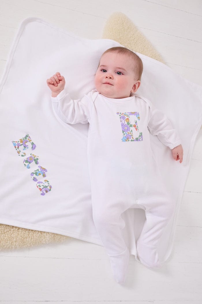 a happy baby wearing a cotton baby romper with her initial on the front in betsy purple floral liberty print. she is laying on a  cotton blanket with her name sewn on.