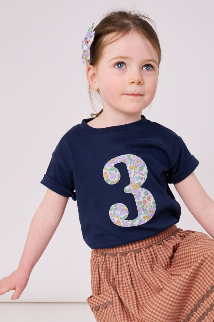 A 3 year old birthday girl wearing a navy cotton kids birthday t-shirt with the  number 3 in Betsy purple floral Liberty print on the front.