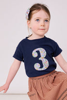 A 3 year old birthday girl wearing a navy cotton kids birthday t-shirt with the  number 3 in Betsy purple floral Liberty print on the front.