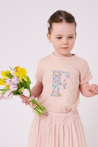 a little girl wearing a personalised kids blush pink  t-shirt with an initial cut from Betsy Purple liberty floral print on the front.
