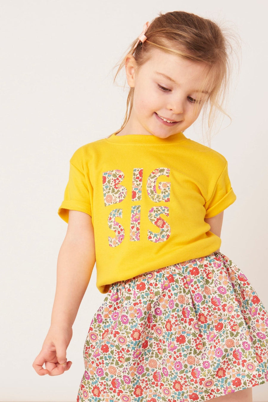 Child wearing a yellow 'BIG SIS' t-shirt with a floral skirt on a white background