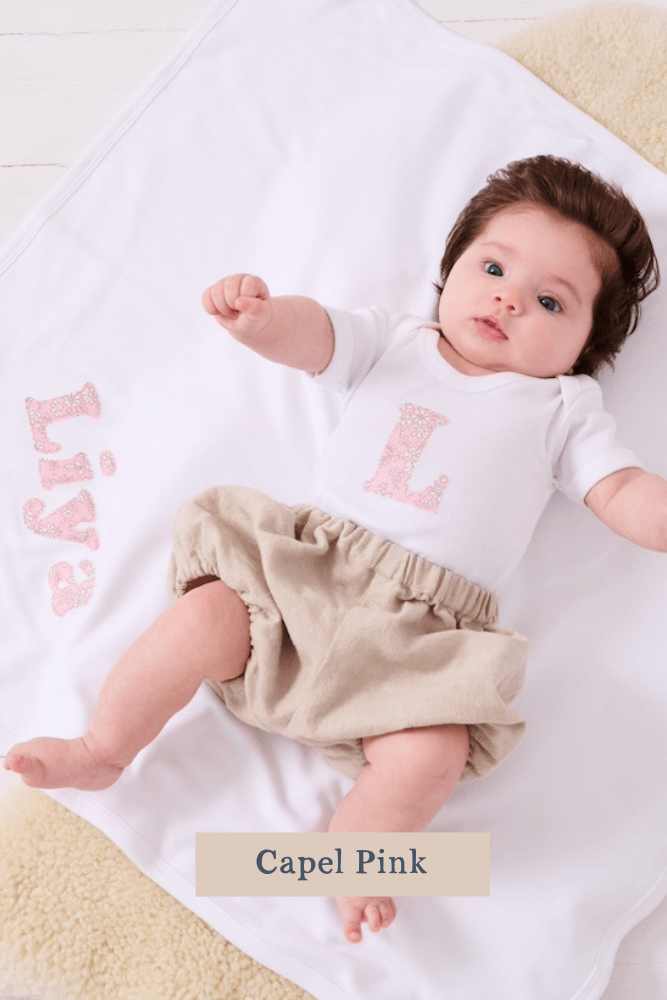 A smiling newborn baby wearing a personalised white bodysuit with an initial on the front cut from Capel Pink Liberty floral fabric. She is laying on a personalised blanket with  her name on.