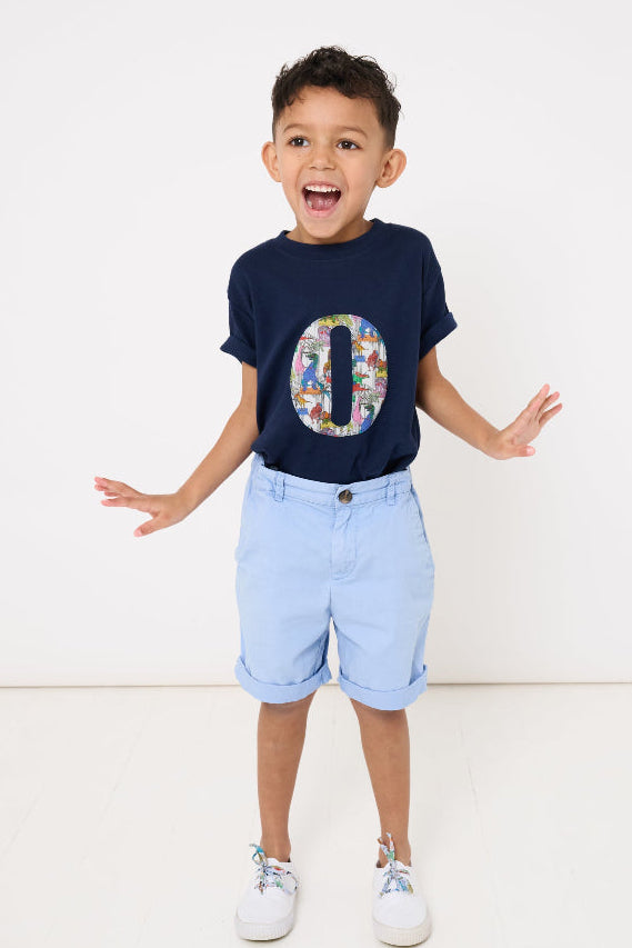 A 4 year old boy wearing a bespoke navy t-shirt with a colourful letter initial design in Liberty dinosaur print and light blue shorts on a white background