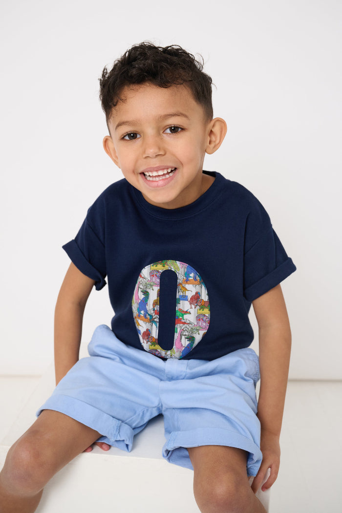 A 4 year old boy wearing a personalised navy t-shirt with a colourful  letter design in Liberty dinosaur print and light blue shorts on a white background