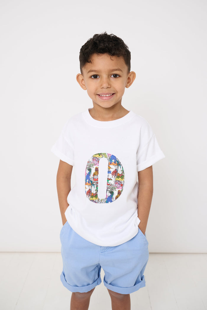 A smiling 4 year old  wearing a white kids T-Shirt with his initial on the front in Dinosaur Liberty Print