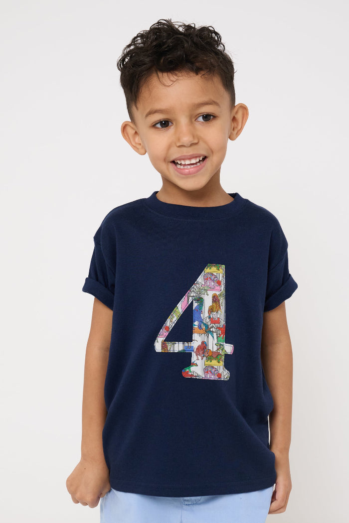 A 4 year old boy wearing a navy t-shirt with a colourful number 4 design in Liberty dinosaur print and light blue shorts on a white background