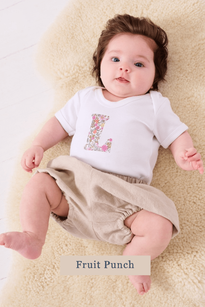 A smiling newborn baby wearing a personalised white bodysuit with an initial on the front cut from Fruit punch Liberty floral fabric.