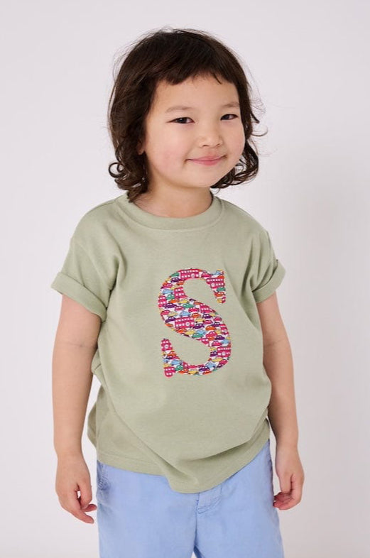 a 4 year old boy wearing a personalised kids sage green t-shirt with an initial cut from hop on hop off res london bus and cars liberty print on the front.