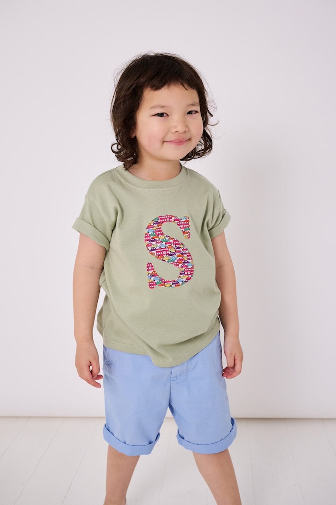 a 4 year old boy wearing a personalised kids sage green t-shirt with an initial cut from hop on hop off red london bus and cars liberty print on the front.