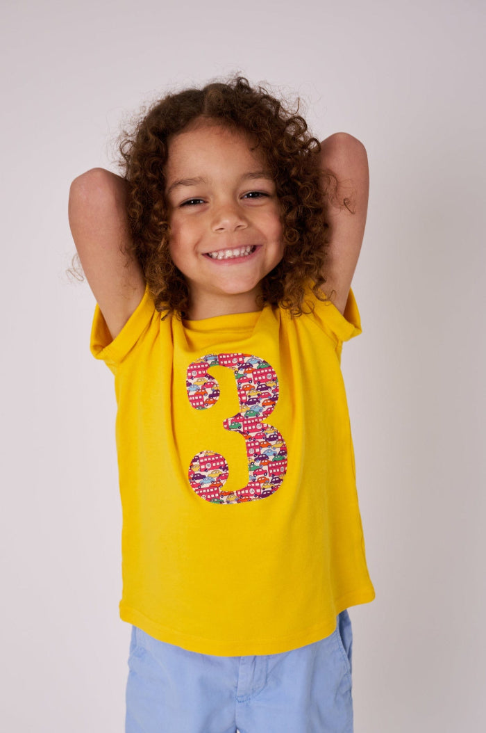 a  3 year old birthday boy wearing a yellow t-shirt with a number 3 cut from Hop on hop off red cars and london bus liberty print on the front.