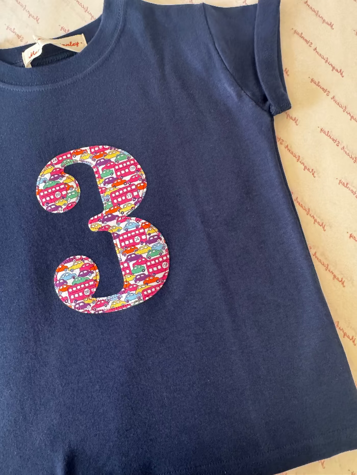 A kids navy blue short-sleeve t-shirt featuring a large, colourful Liberty car print applique number '3' on the front, laid flat on a patterned beige surface.
