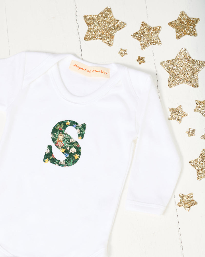 A white long-sleeve baby bodysuit laid flat on a white surface, featuring a decorative capital letter 'S' on the chest with a green Liberty Elves pattern.