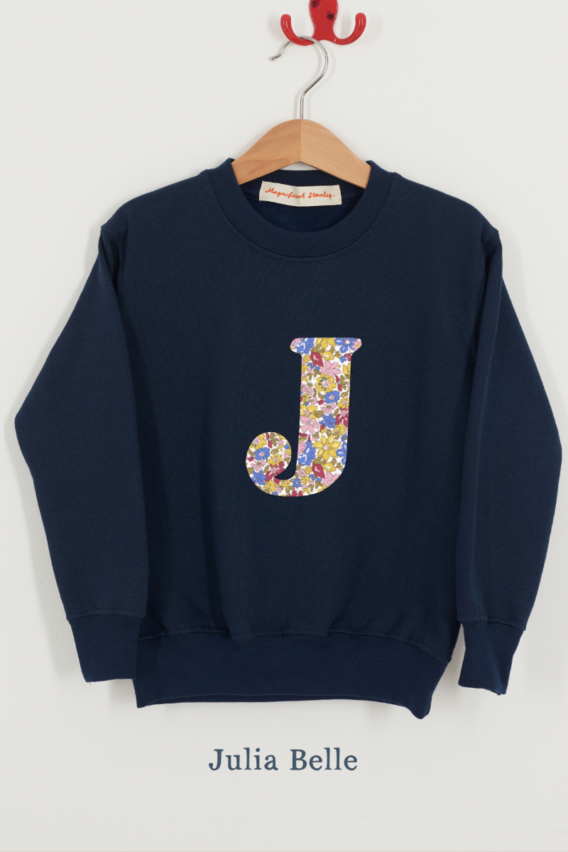 A navy blue crewneck sweatshirt hanging on a wooden hanger against a light gray wall. The sweatshirt features a large, stylized letter 'J' on the chest filled with a Julia Belle Liberty sparkly floral pattern.