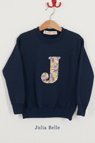 A navy blue crewneck sweatshirt hanging on a wooden hanger against a light gray wall. The sweatshirt features a large, stylized letter 'J' on the chest filled with a Julia Belle Liberty sparkly floral pattern.