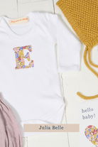 A white long-sleeve baby bodysuit laid flat on a white wood background, featuring a colorful floral monogram letter 'E' on the chest made from Liberty Julia Belle cotton. Styled flat-lay props include a mustard yellow knit hat, a pink fabric item, and a small card reading 'hello baby!'.