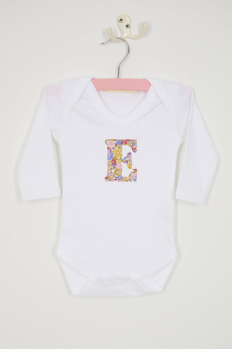 A white long-sleeve baby bodysuit laid flat on a yellow and pink sparkly Liberty Julia Belle floral fabric background. The bodysuit has a decorative floral block-letter 'E' on the chest.
