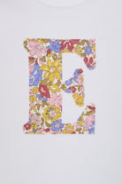 A close up of a white long-sleeve baby bodysuit laid flat on a yellow and pink sparkly Liberty Julia Belle floral fabric background. The bodysuit has a decorative floral block-letter 'E' on the chest.