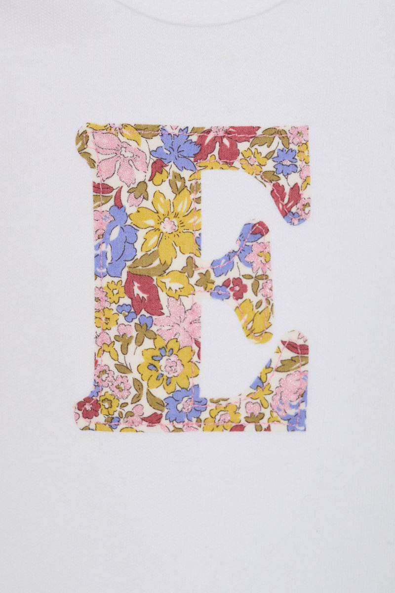 A close up of a white long-sleeve baby bodysuit laid flat on a yellow and pink sparkly Liberty Julia Belle floral fabric background. The bodysuit has a decorative floral block-letter 'E' on the chest.