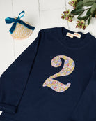 A navy blue long-sleeve baby/toddler t-shirt with a large floral Liberty Julia Belle number '2' on the front, styled on a white wooden background with a pink party hat.