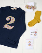 A navy blue long-sleeve baby/toddler t-shirt with a large floral Liberty Julia Belle number '2' on the front, styled on a white wooden background with matching liberty scrunchie and clips