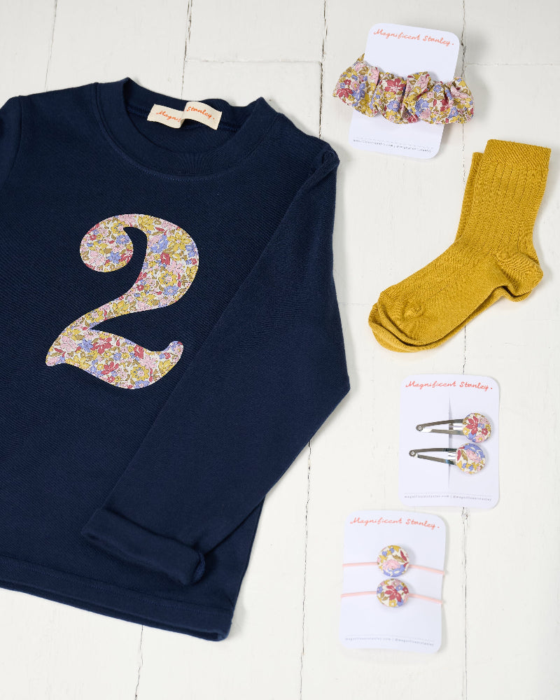 A navy blue long-sleeve baby/toddler t-shirt with a large floral Liberty Julia Belle number '2' on the front, styled on a white wooden background with matching liberty scrunchie and clips