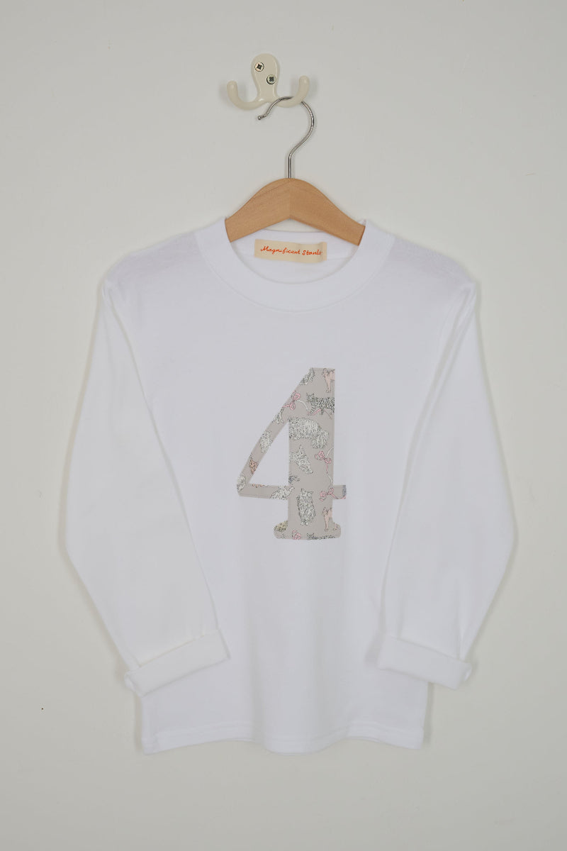 A white long-sleeve t-shirt with a large number 4 printed on the front in a light pink and beige Liberty cat print, hanging on a wooden hanger against a light grey wall.