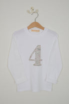 A white long-sleeve t-shirt with a large number 4 printed on the front in a light pink and beige Liberty cat print, hanging on a wooden hanger against a light grey wall.
