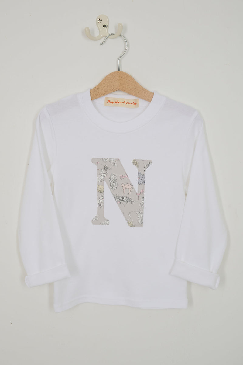 A white long-sleeve t-shirt for kids, hanging on a wooden hanger. The shirt features a large decorative letter 'N' on the front filled with a Liberty Koh & Eito cats print in muted gray, beige, and pink tones.