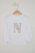 A white long-sleeve t-shirt for kids, hanging on a wooden hanger. The shirt features a large decorative letter 'N' on the front filled with a Liberty Koh & Eito cats print in muted gray, beige, and pink tones.
