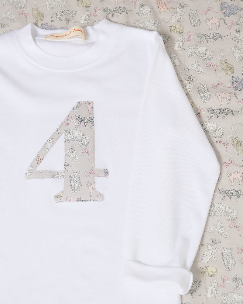 A children's outfit styled flat-lay on a white wood background, featuring a white long-sleeve top with a large decorative number 4 on the front in Liberty Koh Eito cats print .