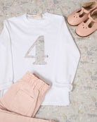 A children's outfit styled flat-lay on a white wood background, featuring a white long-sleeve top with a large decorative number 4 on the front in Liberty Koh Eito cats print.