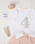 A children's outfit styled flat-lay on a white wood background, featuring a white long-sleeve top with a large decorative number 4 on the front in Liberty Koh Eito cats print and a light pink tulle skirt. 