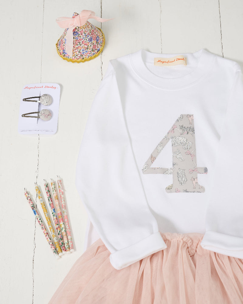 A children's outfit styled flat-lay on a white wood background, featuring a white long-sleeve top with a large decorative number 4 on the front in Liberty Koh Eito cats print and a light pink tulle skirt. 