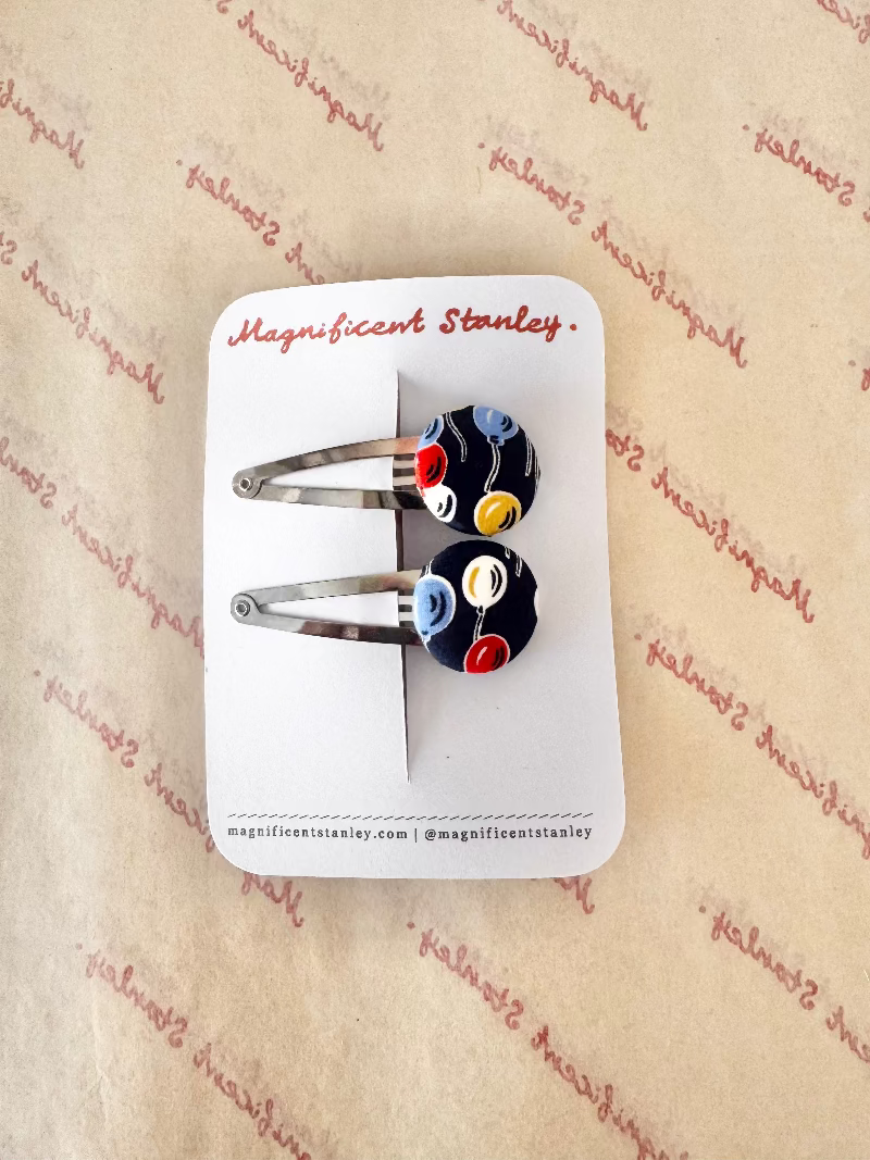 A pair of silver metal hair clips with colorful glass bead charms, displayed on a white branded card that reads 'Magnificent Stanley'. The card is placed on a beige background with a repeating red script pattern.